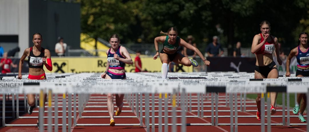 Drei weibliche Athleten springen über Hürden auf einer Laufbahn. Eine führt mit Startnummer 182, gefolgt von 413, und eine weitere im Hintergrund. Zuschauer beobachten von hinten.