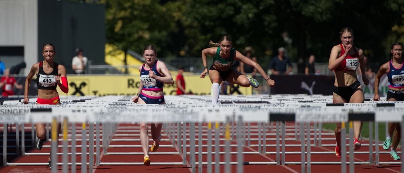 Drei weibliche Athleten springen über Hürden auf einer Laufbahn. Eine führt mit Startnummer 182, gefolgt von 413, und eine weitere im Hintergrund. Zuschauer beobachten von hinten.