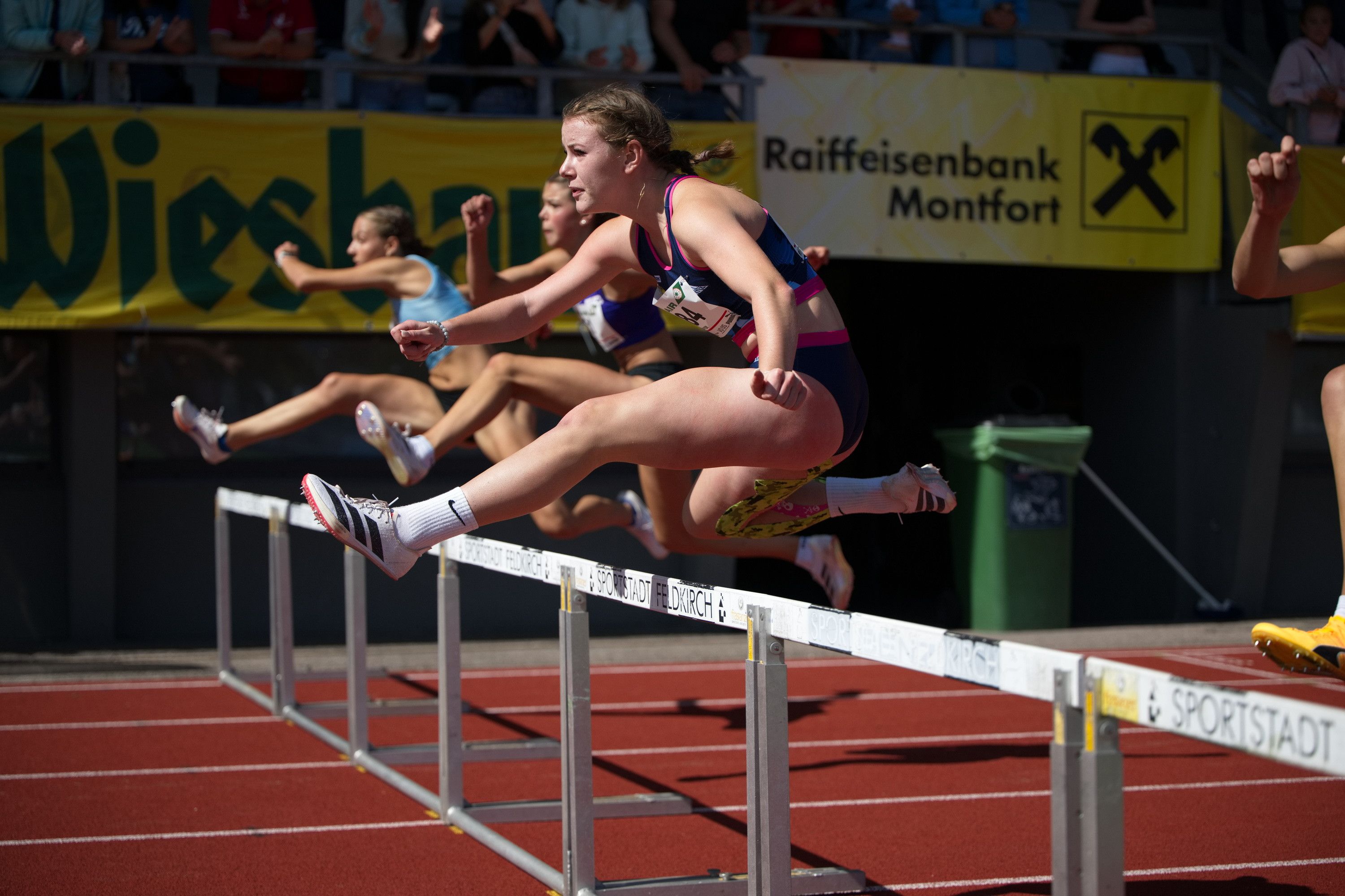 Eine Athletin bei einem Leichtathletik-Wettkampf springt über Hürden auf einer roten Laufbahn. Hinter ihr konkurrieren andere Athleten. Ein Schild trägt die Aufschrift Raiffeisenbank Montfort.