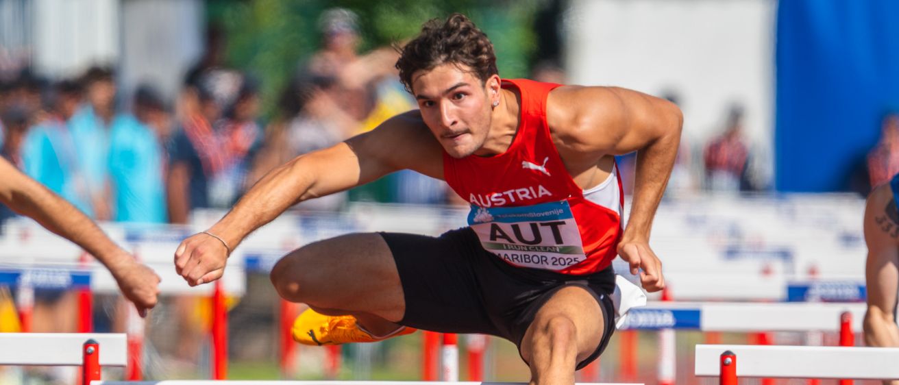 Ein männlicher Athlet aus Österreich springt auf einer Laufbahn über Hürden und trägt rote und weiße Sportkleidung.
