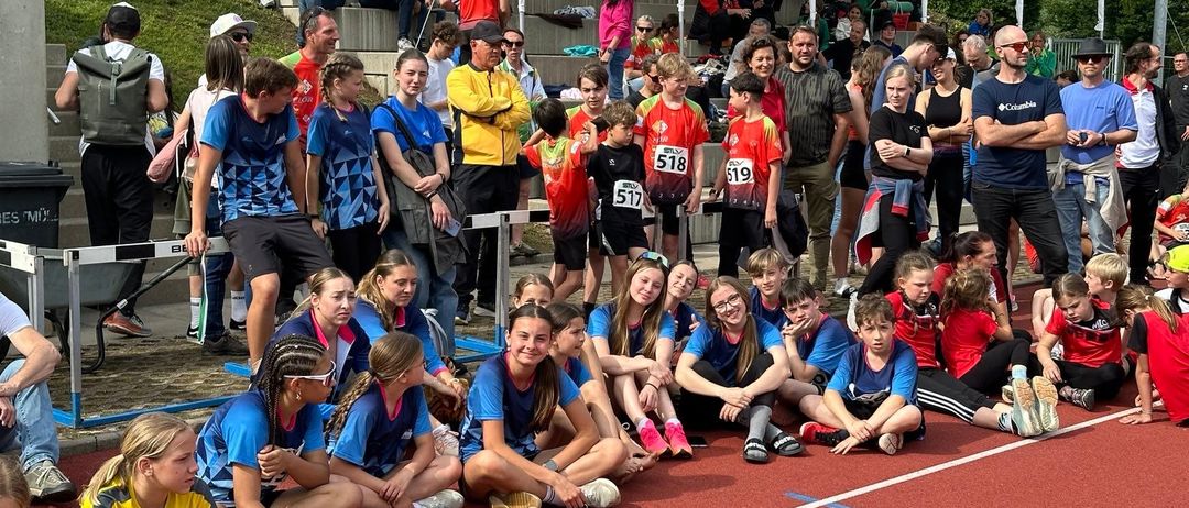 Eine Gruppe von Athleten in blauen Trikots sitzt auf der Laufbahn. Zuschauer in lässiger Kleidung sitzen auf den Tribünen. Die Atmosphäre ist lebhaft, während alle auf den Beginn der Veranstaltung warten.