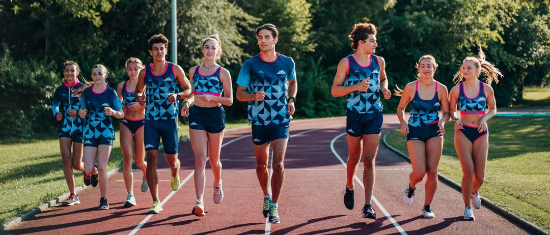 Eine Gruppe junger Athleten läuft auf einer Laufbahn mit Bäumen und einem Pfahl im Hintergrund. Sie tragen passende blaue Uniformen mit geometrischen Mustern.
