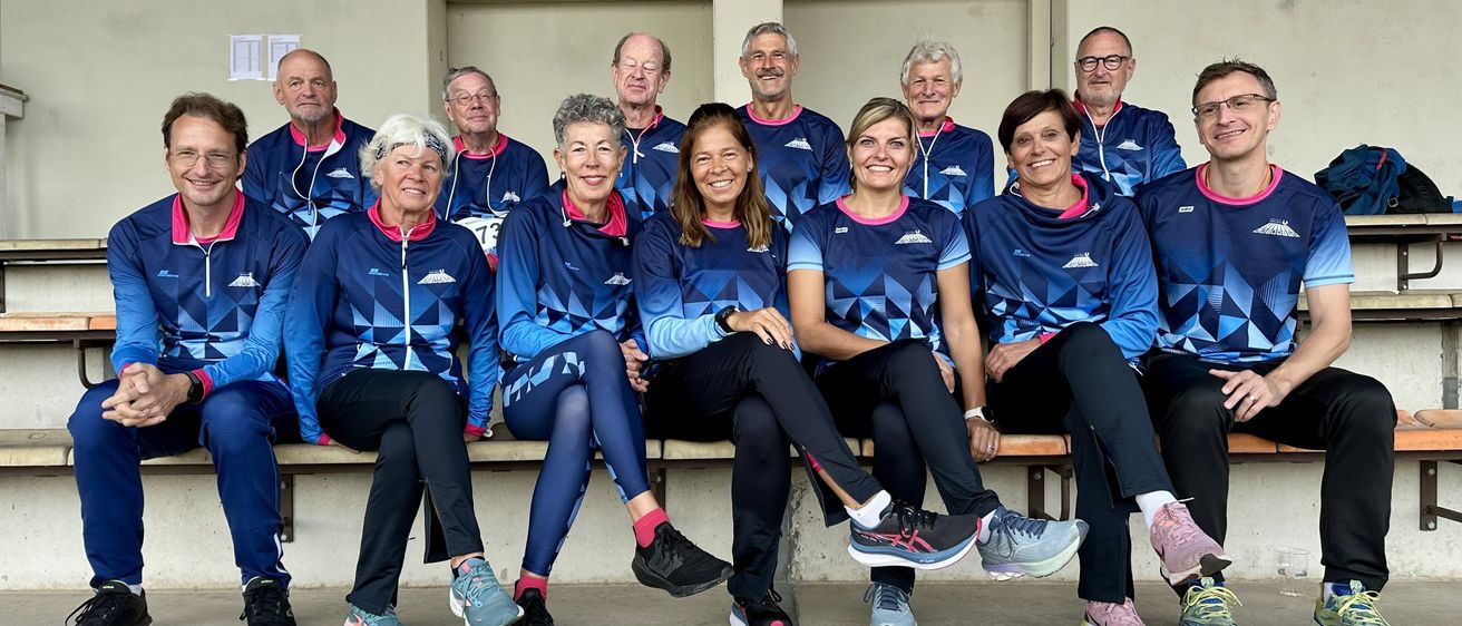 Eine Gruppe von Erwachsenen in passenden blauen und rosafarbenen Uniformen posiert für ein Foto auf einer Bank. Sie lächeln und scheinen sich bei einer Sportveranstaltung zu befinden.