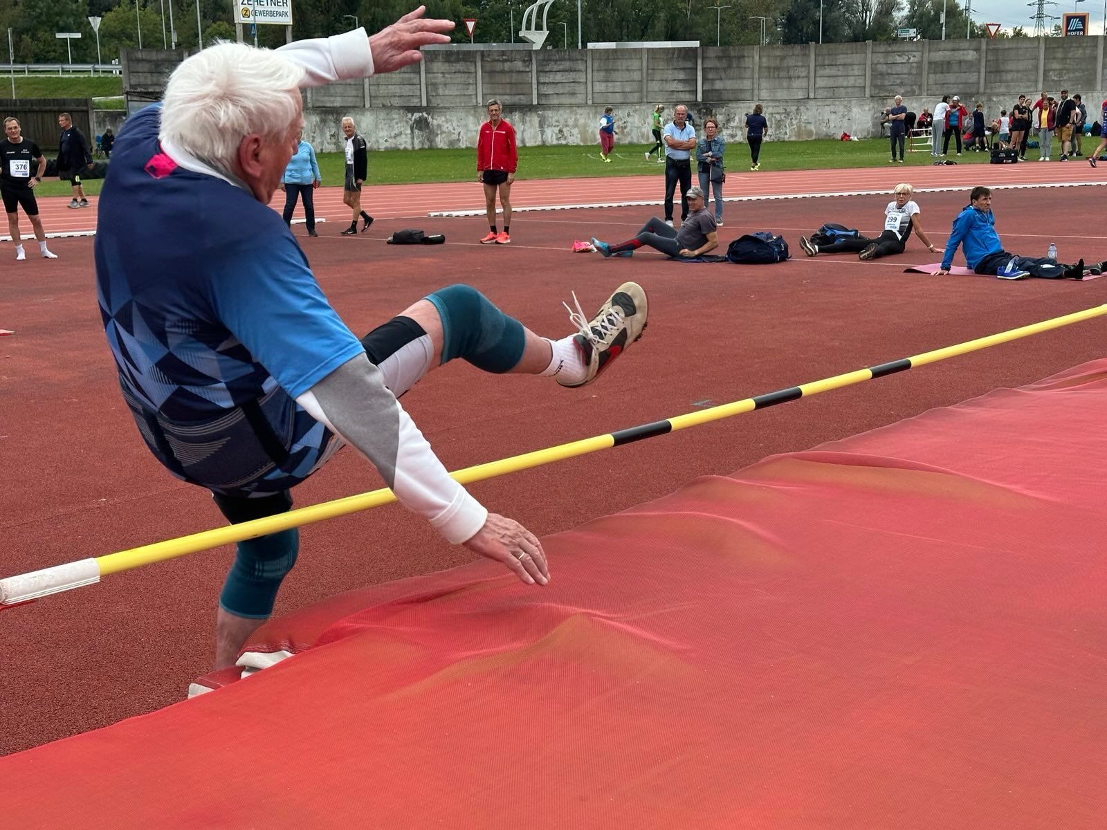 Ein älterer Mann springt über eine gelbe Stange bei einem Leichtathletik-Event. Zuschauer sind auf der Laufbahn verteilt.
