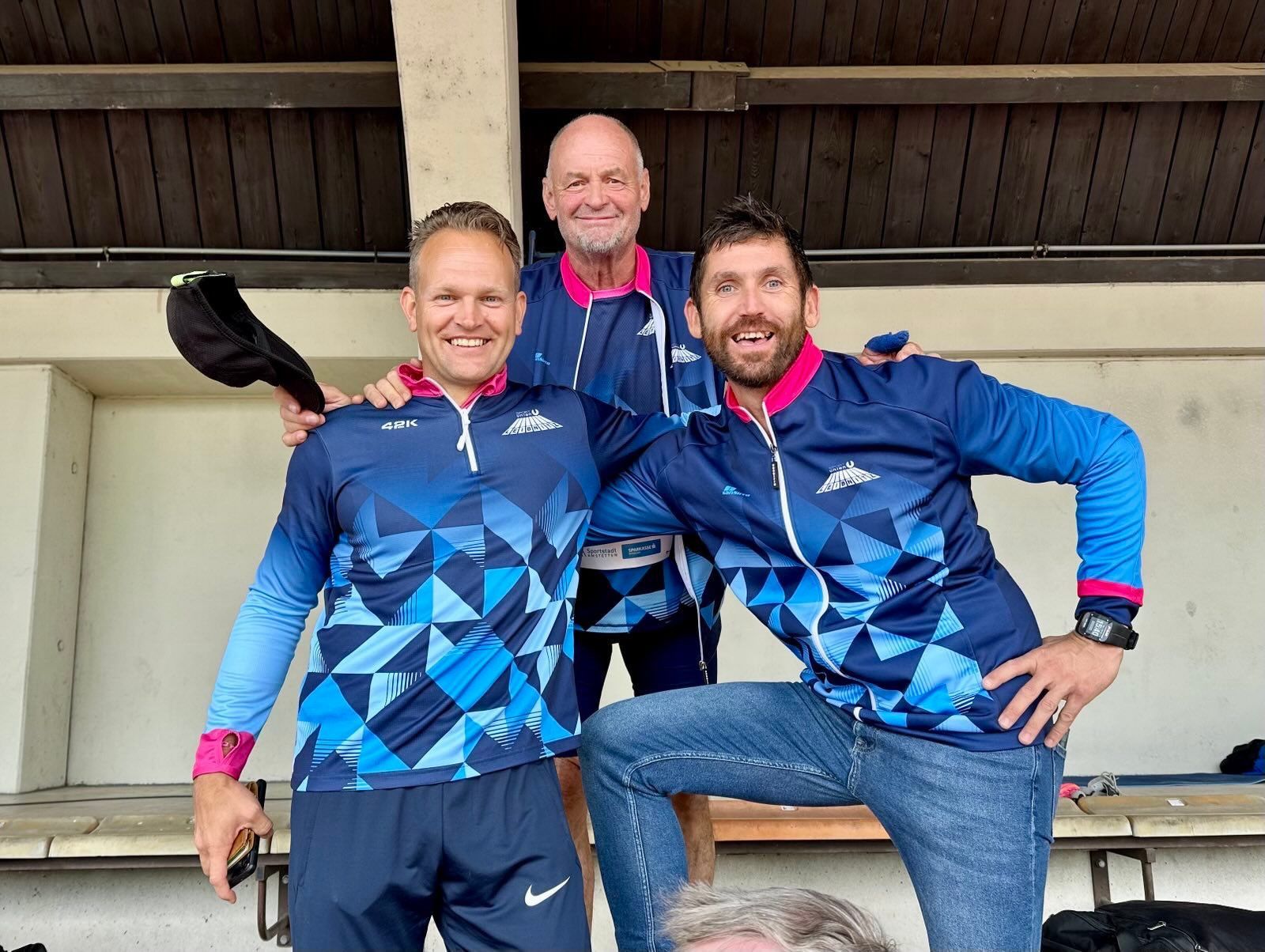 Drei Männer in blauer und rosa Sportbekleidung posieren für ein Foto. Zwei stehen, während der dritte seine Arme um die Schultern der anderen gelegt hat. Sie alle haben breite Lächeln.