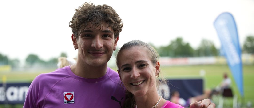 Ein Mann und eine Frau stehen zusammen und lächeln für ein Foto bei einer Sportveranstaltung. Beide tragen pinkfarbene Shirts mit weißen Logos. Im Hintergrund sind verschwommen Menschen und eine Rasenfläche zu sehen.