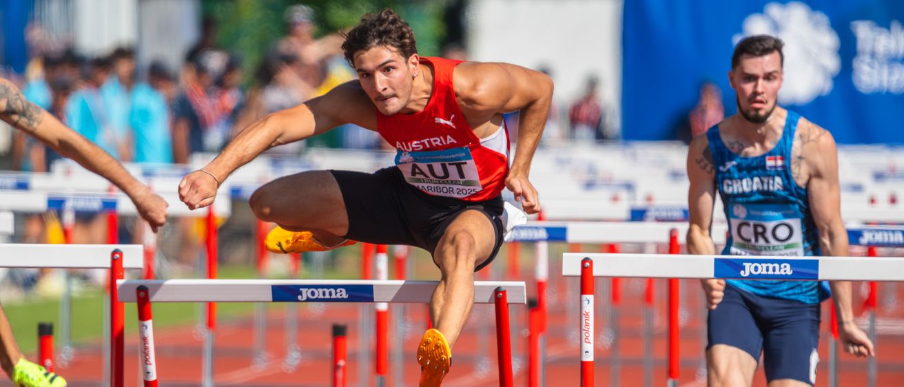 Ein männlicher Athlet springt über eine Hürde bei einem Leichtathletikwettbewerb. Er trägt ein rotes Österreich-Trikot und orangefarbene Schuhe. Ein anderer Läufer folgt ihm.