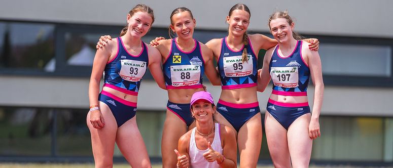 Fünf weibliche Athletinnen stehen auf einer Laufbahn. Sie tragen blaue und rosa Uniformen mit den Nummern 93, 88, 95, 197 und eine Frau kniet in einem weißen Tanktop. Sie scheinen zu feiern.
