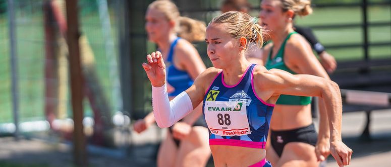 Eine Läuferin mit der Nummer 88 auf ihrem Trikot sprintet auf einer Laufbahn, mit anderen Läuferinnen hinter ihr. Sie trägt ein blaues und rosafarbenes Sportoutfit mit der Marke Eva Air.