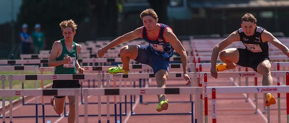 Athleten springen über Hürden bei einem Leichtathletikwettbewerb. Vier Athleten in blauen und schwarzen Uniformen konkurrieren auf einer roten Laufbahn.