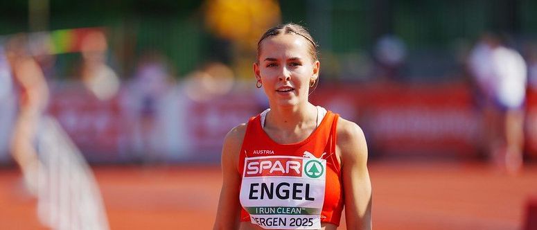 Eine Athletin in einem orangefarbenen Trikot mit dem Namen 'Engel' läuft auf einer Laufbahn, möglicherweise während eines Wettkampfs. Der Hintergrund ist unscharf, mit sichtbaren Personen und Barrieren.