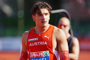 Ein Mann in einem orangefarbenen Singlet mit dem Aufdruck Austria läuft auf einer Laufbahn. Er trägt eine Halskette und rote Armbänder. Im unscharfen Hintergrund sitzt ein Mann auf einem Stuhl.