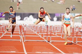 Drei Athletinnen konkurrieren in einem Hürdenrennen. Die erste Athletin springt, während die zweite in der Luft ist. Die dritte Athletin läuft auf der Laufbahn.