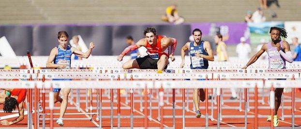 Athleten konkurrieren in einem Hürdenrennen auf einer Laufbahn, mit Hürden zwischen ihnen. Zuschauer sitzen im Hintergrund.
