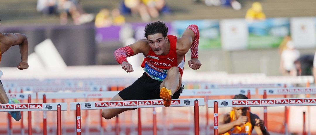 Drei Athleten springen über Hürden auf einer Laufbahn, einer ist in der Luft. Zuschauer sitzen im Hintergrund. Der Athlet in der Mitte trägt ein rotes Trikot mit dem Text Austria. Der Athlet auf der linken Seite trägt ein orangefarbenes Trikot mit dem Text Germany. Der Athlet auf der rechten Seite trägt ein orangefarbenes Trikot mit dem Text Sophia.