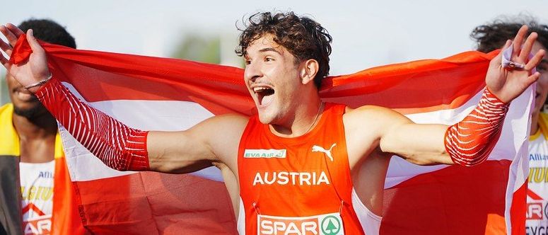 Ein österreichischer Läufer hält eine Flagge in der Hand, während er einen Marathon läuft, und trägt ein orangefarbenes Trikot mit dem Wort Österreich und dem Namen Diesel.