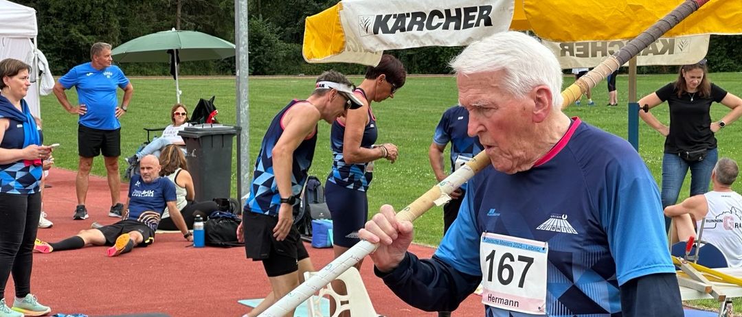 Ein älterer Mann mit der Nummer 167 hält eine Stabhochsprungstange. Hinter ihm beugt sich eine Person. Ein weißer Stuhl und ein Zelt mit dem Wort 'Karcher' sind in der Nähe.