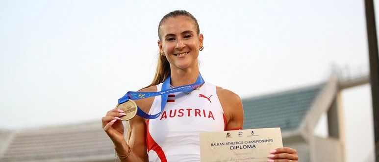 Eine Athletin mit einer österreichischen Medaille und Diplom posiert lächelnd bei einer Veranstaltung im Freien mit einem unscharfen Stadionhintergrund.