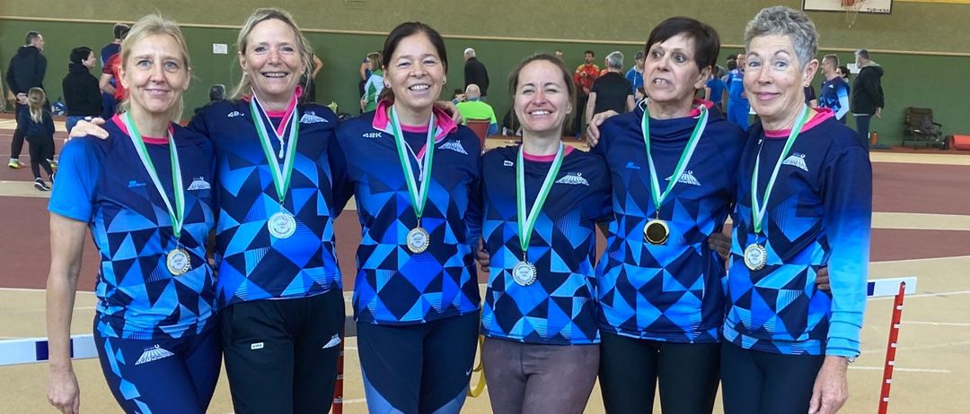 Fünf Frauen in blauen Uniformen mit Medaillen um den Hals stehen in einer Reihe, lächeln in die Kamera, in einer Turnhalle.
