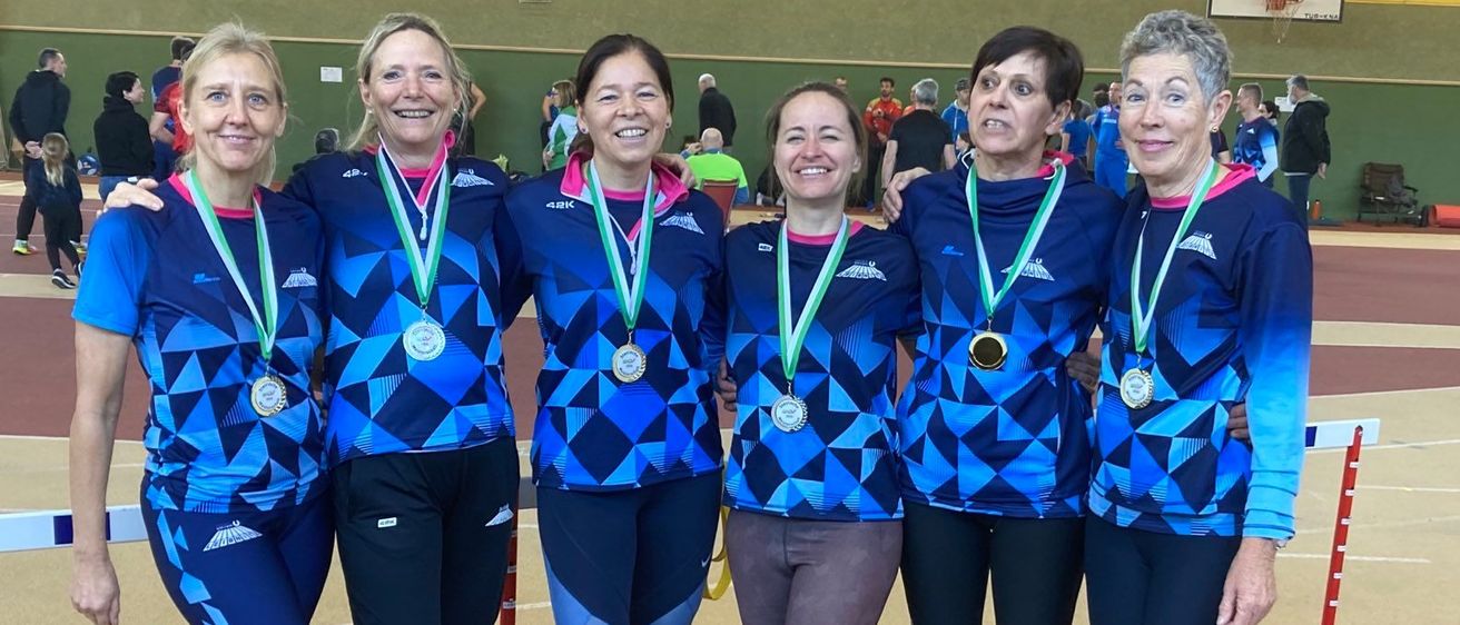 Fünf Frauen in blauen Uniformen mit Medaillen um den Hals stehen in einer Reihe, lächeln in die Kamera, in einer Turnhalle.