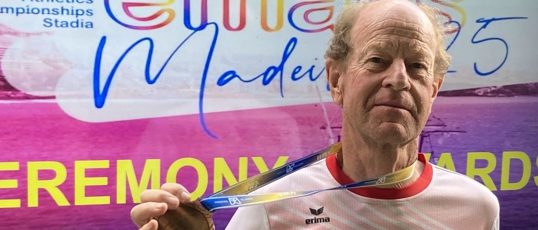 Ein älterer Mann hält eine Goldmedaille mit 'Austria' auf seinem Shirt vor einem farbenfrohen Schild.