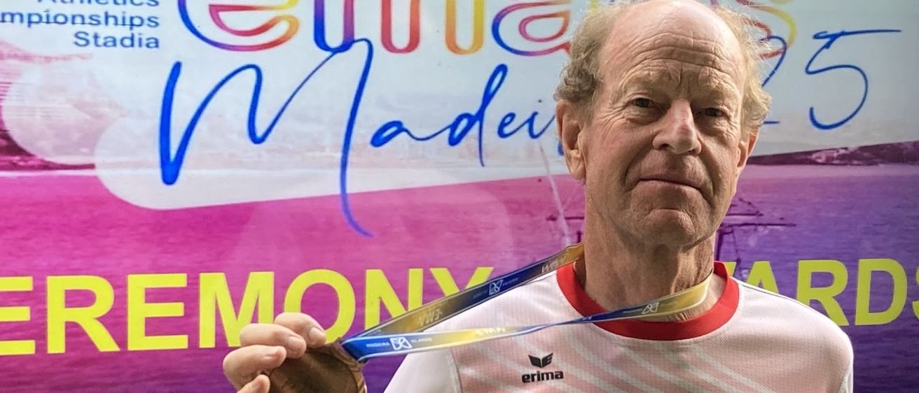 Ein älterer Mann hält eine Goldmedaille mit 'Austria' auf seinem Shirt vor einem farbenfrohen Schild.