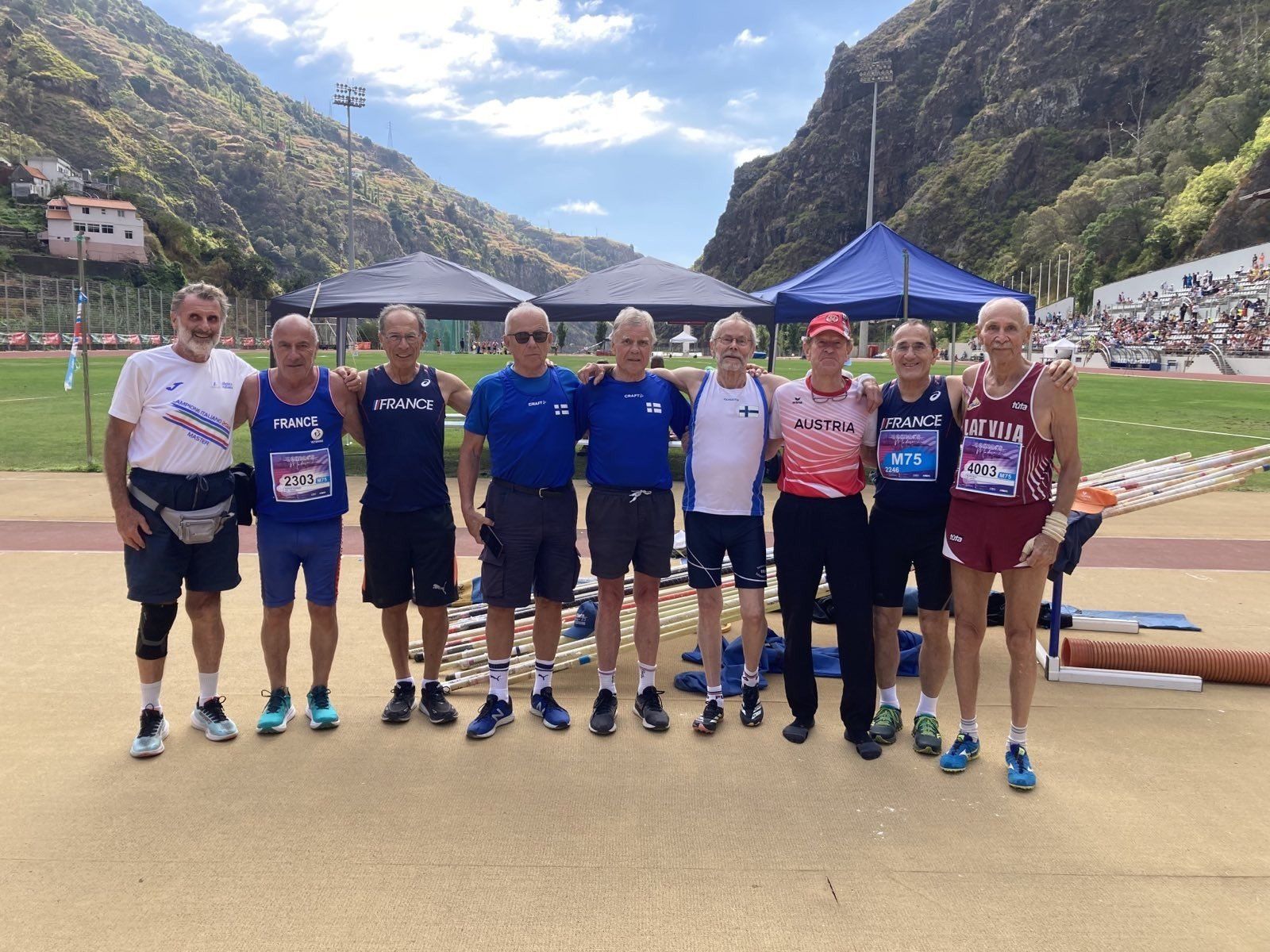 Eine Gruppe älterer Männer in Sportbekleidung und Startnummern posiert für ein Foto auf einer Freiluftlaufbahn mit Bergen im Hintergrund.