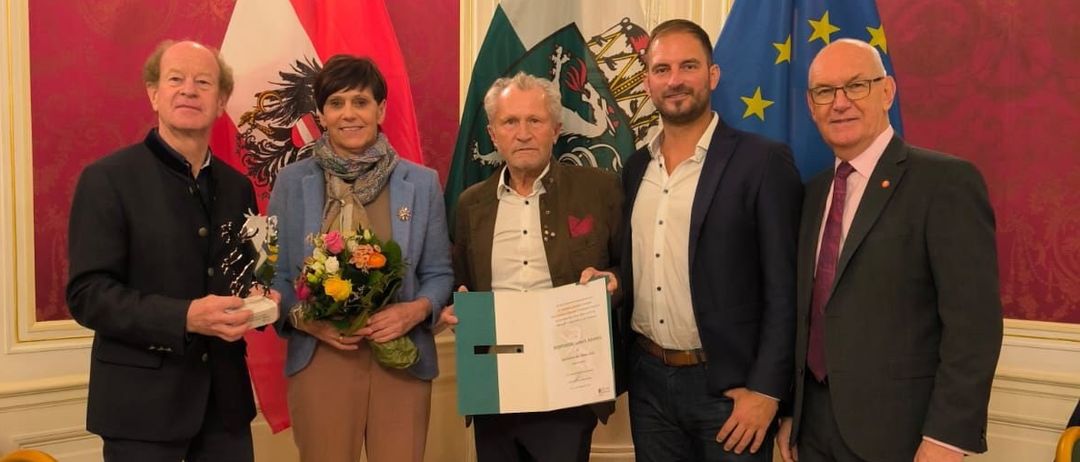 Fünf Personen stehen in einem formellen Ambiente, einer Mann hält ein Zertifikat. Hinter ihnen sind die Flaggen von Österreich, einem Mitglied der Europäischen Union, und eines anderen Landes zu sehen.