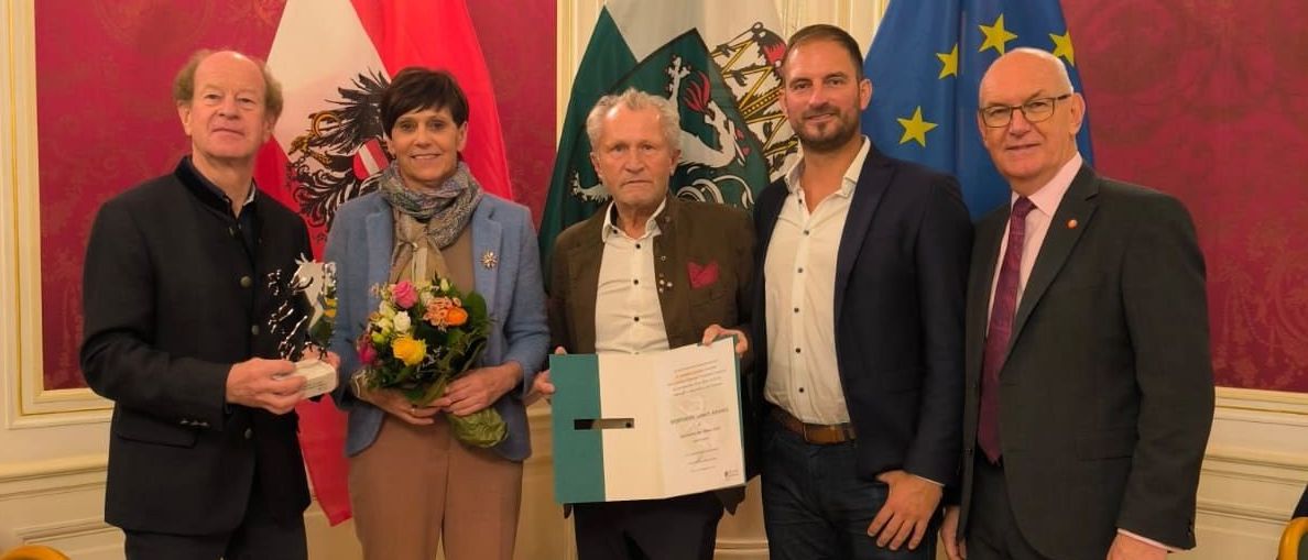 Fünf Personen stehen in einem formellen Ambiente, einer Mann hält ein Zertifikat. Hinter ihnen sind die Flaggen von Österreich, einem Mitglied der Europäischen Union, und eines anderen Landes zu sehen.