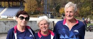 Drei Personen in übereinstimmender blauer Sportbekleidung stehen zusammen, eine hält eine rosa Jacke, in einer Außenumgebung mit einem Hintergrund eines leeren Stadions.