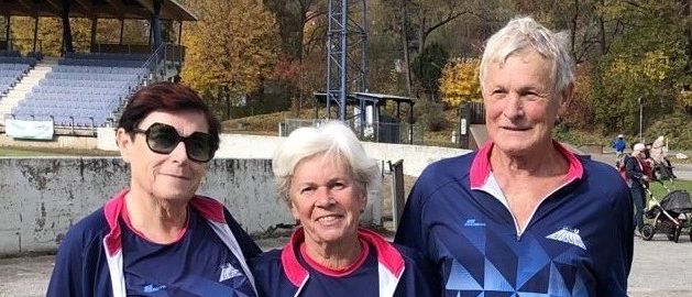Drei Personen in übereinstimmender blauer Sportbekleidung stehen zusammen, eine hält eine rosa Jacke, in einer Außenumgebung mit einem Hintergrund eines leeren Stadions.