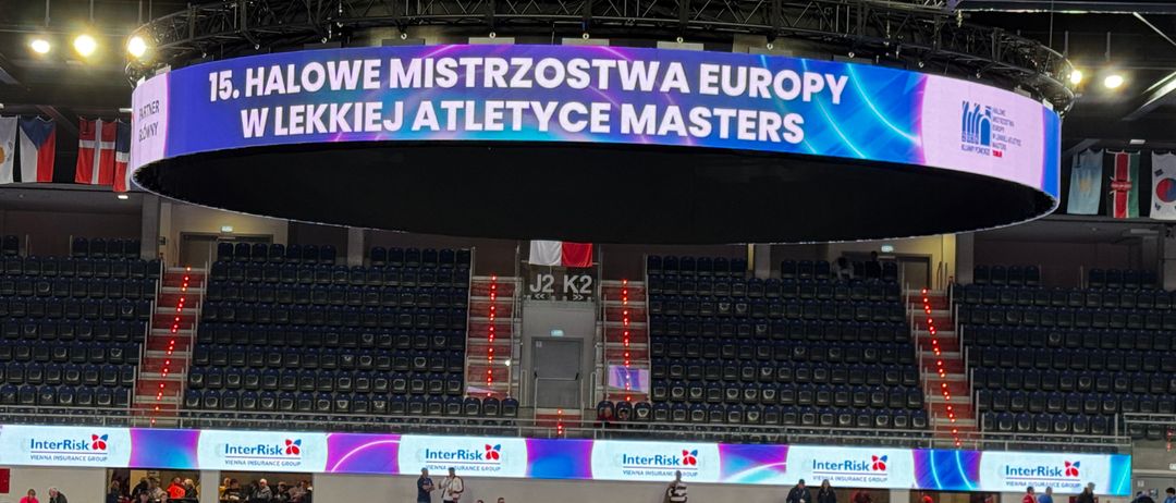 Eine überdachte Sportarena veranstaltet die 15. Halwe Mistrzostwa Europy W Lekcji Atletyce Masters. Leere Sitze umgeben das zentrale Banner, mit Menschen auf den Treppen und einer blau beleuchteten Decke. Werbetafeln säumen die Seiten.