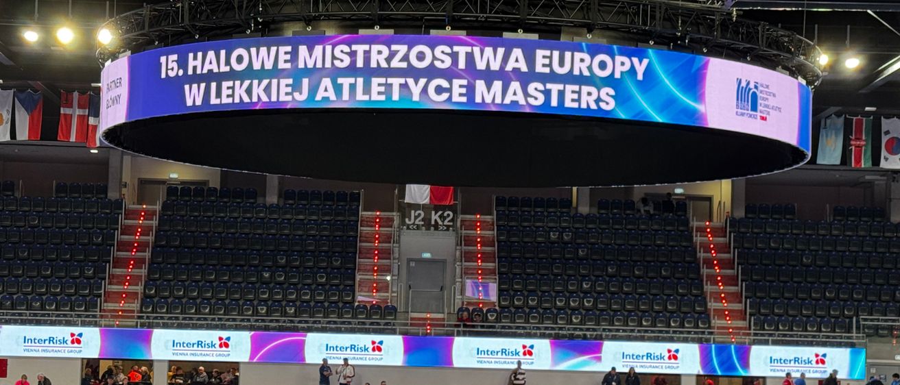 Eine überdachte Sportarena veranstaltet die 15. Halwe Mistrzostwa Europy W Lekcji Atletyce Masters. Leere Sitze umgeben das zentrale Banner, mit Menschen auf den Treppen und einer blau beleuchteten Decke. Werbetafeln säumen die Seiten.