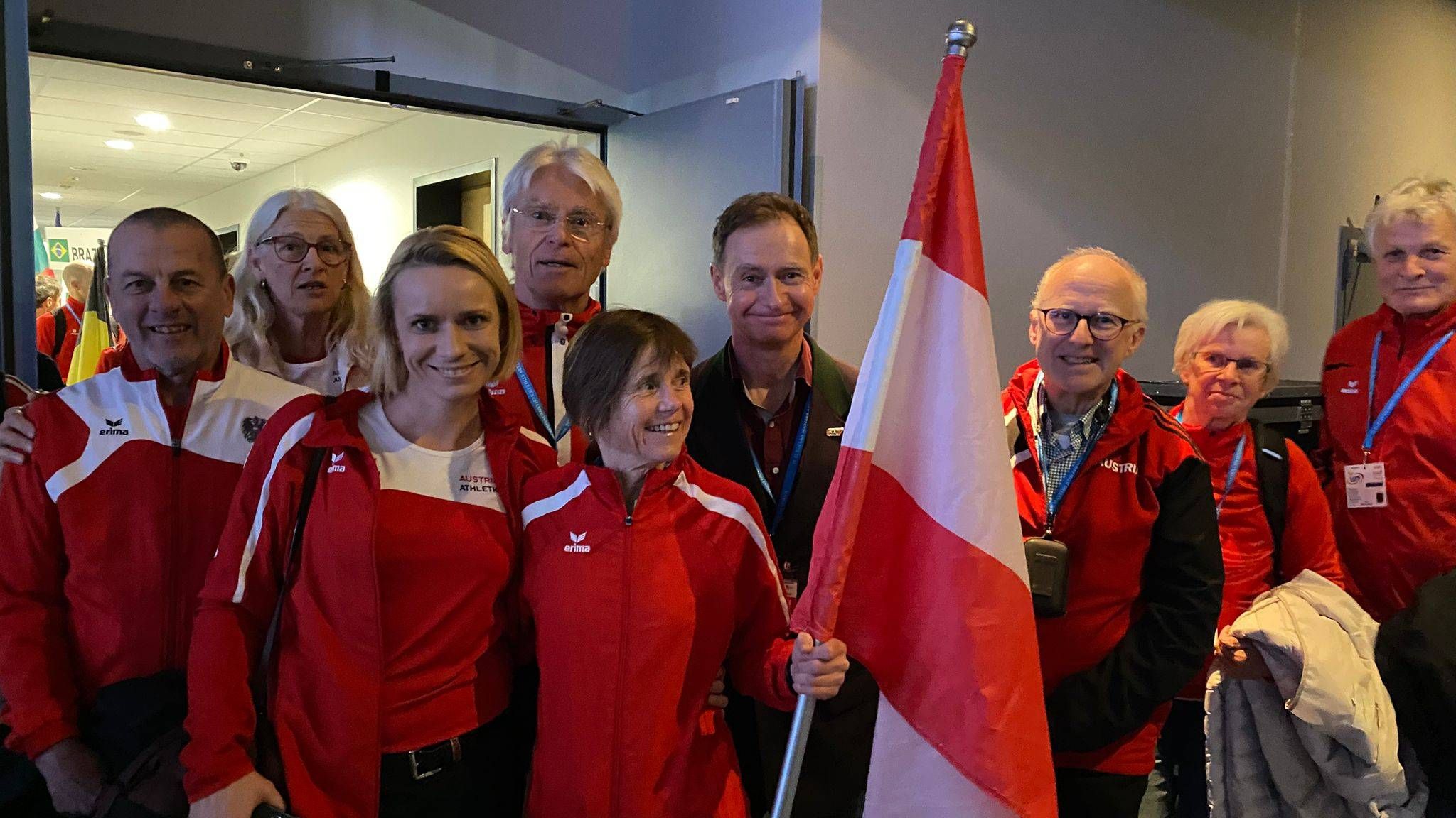 Eine Gruppe von Athleten in roten Sportjacken posiert mit einer Flagge für ein Foto. Sie lächeln und scheinen einen Sieg zu feiern.