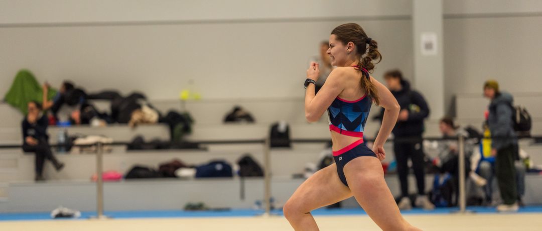Eine Athletin sprintet auf einer Indoor-Laufbahn mit blauen und weißen Markierungen. Sie trägt einen blauen und rosafarbenen Sport-BH und weiße Turnschuhe. Im Hintergrund sind Zuschauer auf den Tribünen.