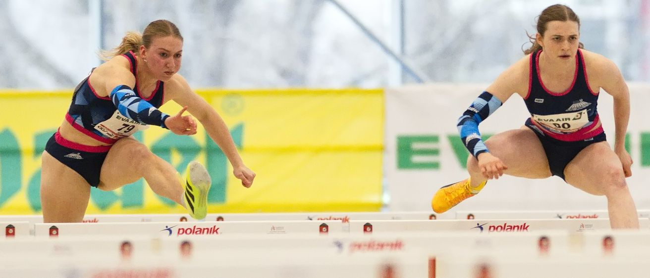 Drei weibliche Athletinnen nehmen an einem Hürdenrennen teil. Sie springen entschlossen über Hindernisse. Die führende Athletin hat die Nummer 90 auf ihrem Trikot.