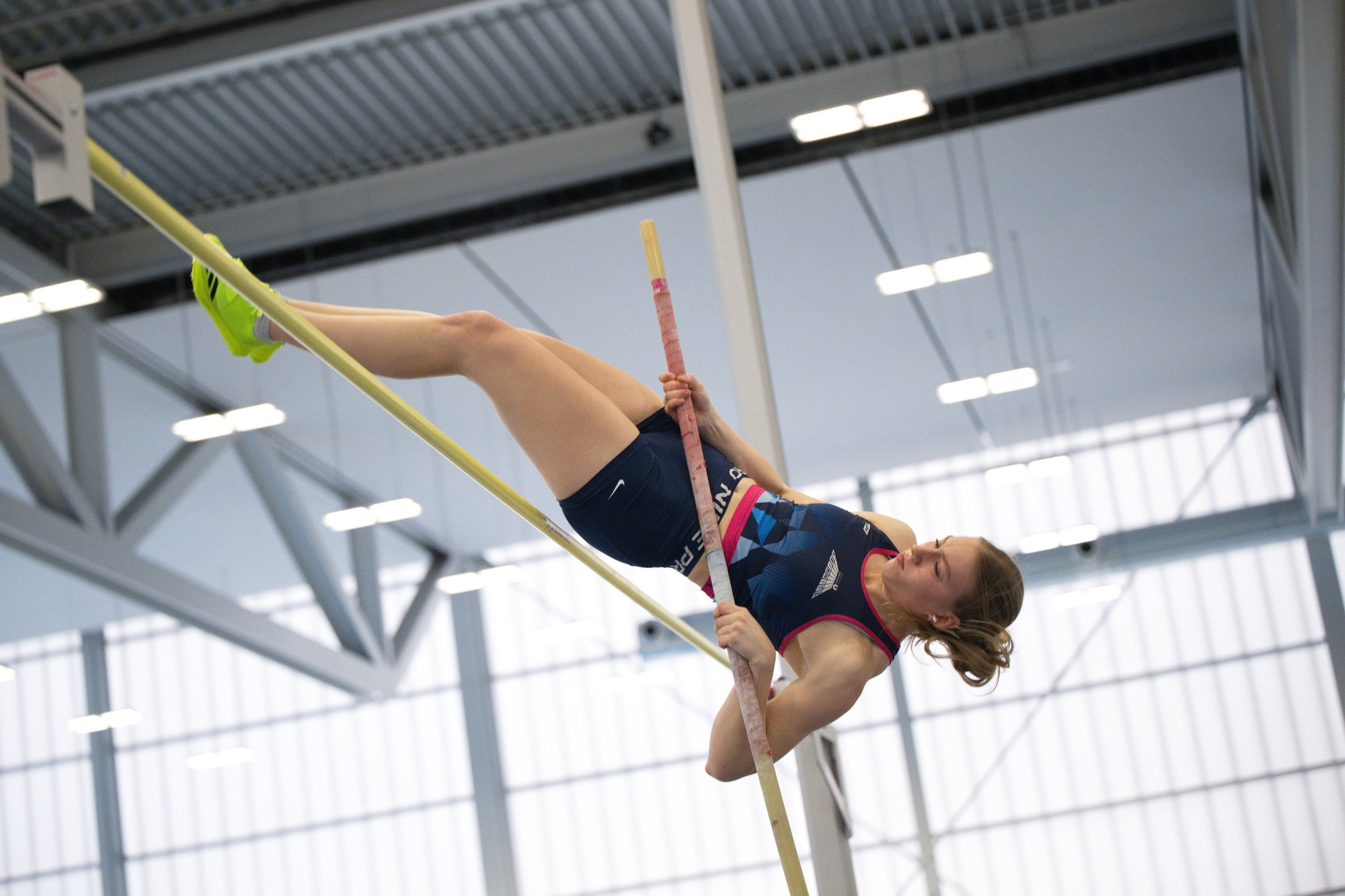 Eine Frau führt einen Stabhochsprung in einer Indoor-Sporthalle aus und hält den Stab mit den Händen fest.