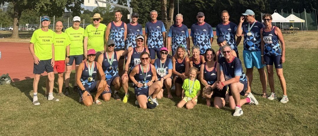 Eine Gruppe von Menschen in passenden blauen und gelben Uniformen posiert für ein Foto auf einem Rasenfeld. Einige tragen Medaillen und Sonnenbrillen. Ein Basketballkorb ist im Hintergrund.