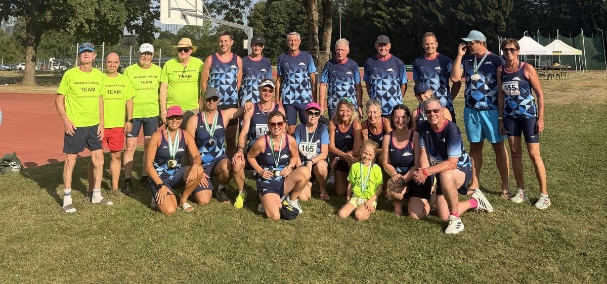 Eine Gruppe von Menschen in passenden blauen und gelben Uniformen posiert für ein Foto auf einem Rasenfeld. Einige tragen Medaillen und Sonnenbrillen. Ein Basketballkorb ist im Hintergrund.