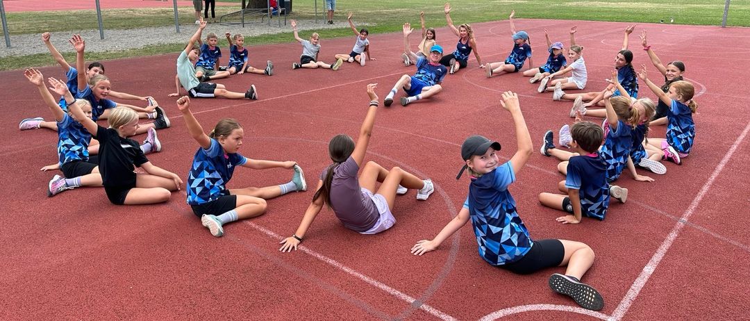 Eine Gruppe von Kindern in blauen und weißen Uniformen macht Aufwärmübungen auf einer roten Laufbahn, während einige Erwachsene in der Nähe stehen.