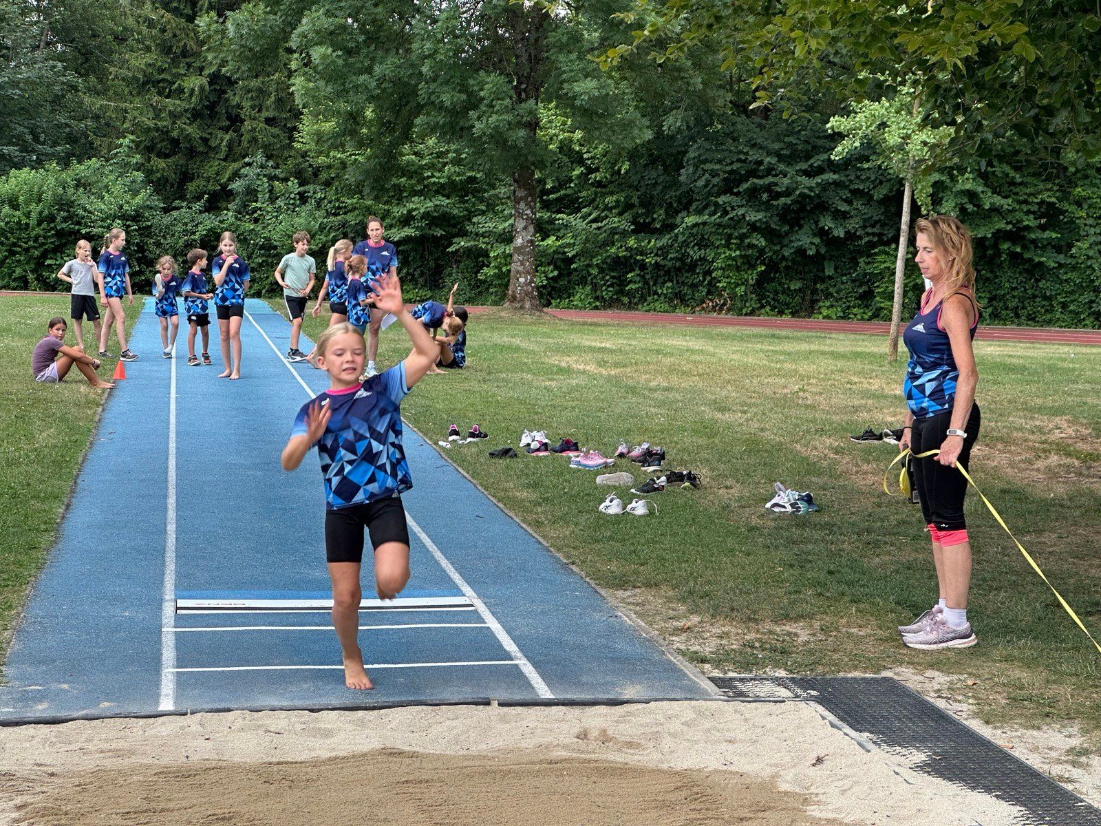 Ein junges Mädchen in einem blau-weißen Trikot springt auf einer Laufbahn, mit anderen Kindern und Erwachsenen in der Nähe.