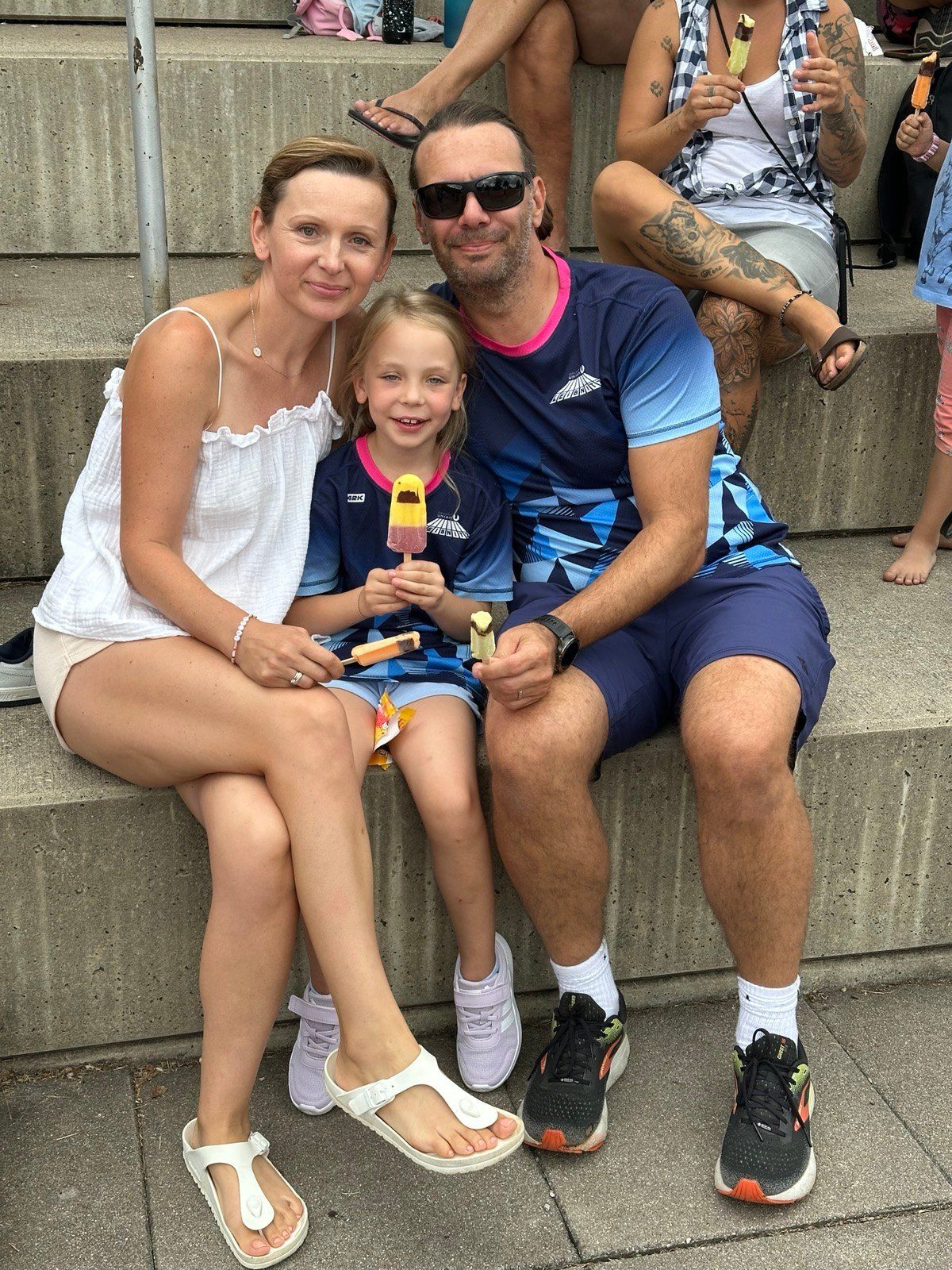Eine Familie aus drei Personen, eine Frau, ein Mann und ein junges Mädchen, sitzen auf einer Betonbank, lächeln und halten Eis am Stiel.