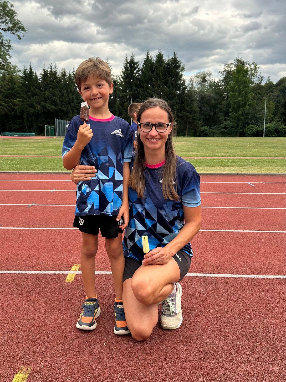 Eine Frau und ein Junge stehen auf einer Laufbahn, beide halten Eis-Popsicles. Der Junge trägt ein blaues Shirt mit geometrischem Muster, und die Frau trägt eine Brille und ein blaues Shirt. Hinter ihnen befindet sich ein Rasenfeld mit Bäumen und einem bewölkten Himmel.