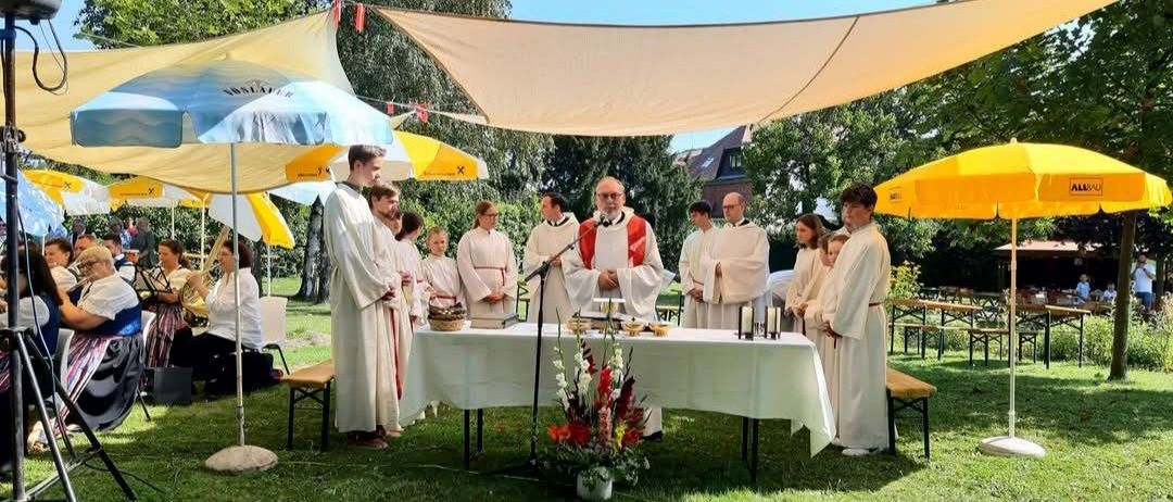 Eine religiöse Zeremonie findet im Freien statt, bei der ein Priester in ein Mikrofon spricht, umgeben von Menschen in weißen Gewändern.