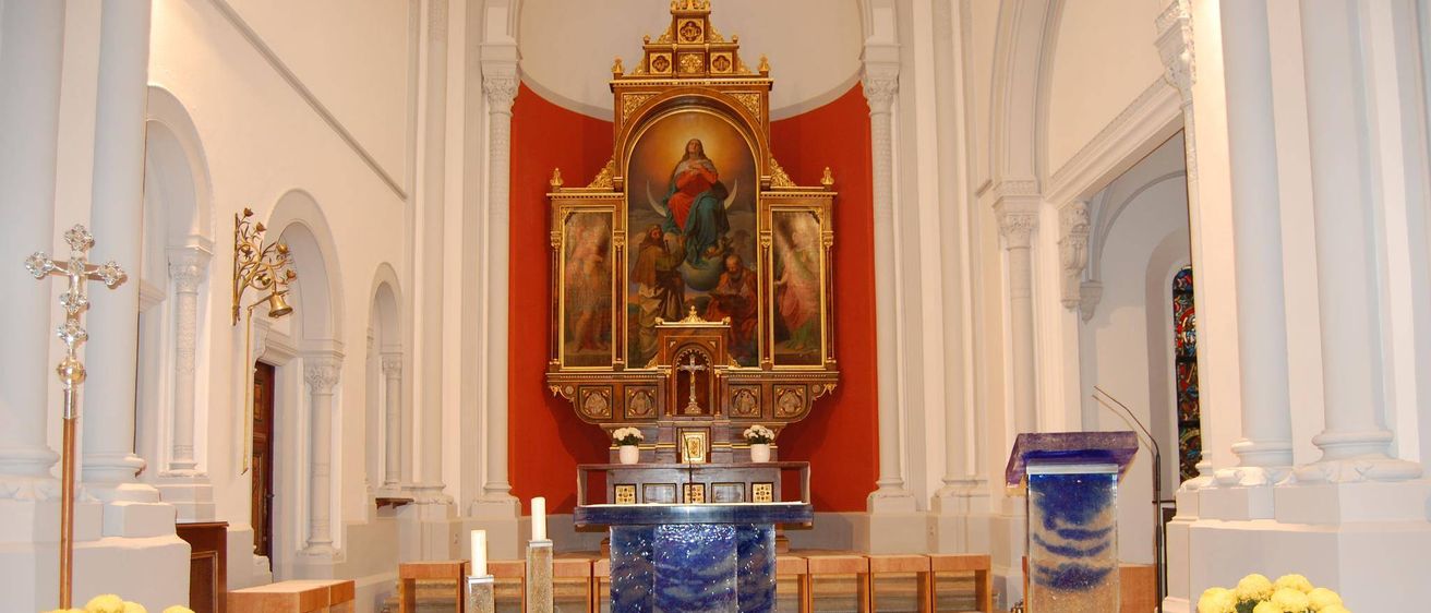 Ein Kirchenaltar mit einem blauen Tisch, einem goldenen Rahmen mit einem religiösen Gemälde, zwei Kerzen und einem Mikrofon auf einem Ständer.