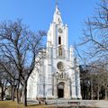 Eine weiße Kirche mit Turm, Uhr und Statuen steht in einem Park mit Bäumen und Bänken unter einem klaren blauen Himmel.