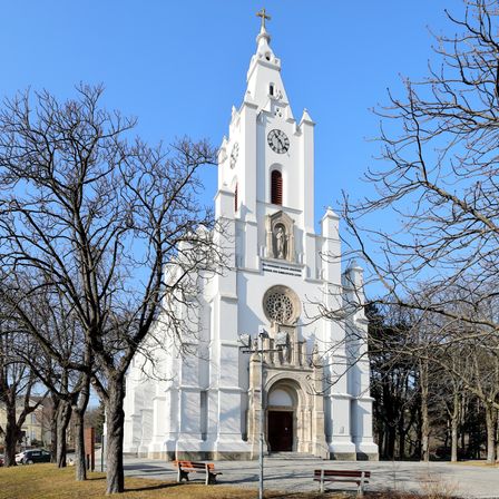 Eine weiße Kirche mit Turm, Uhr und Statuen steht in einem Park mit Bäumen und Bänken unter einem klaren blauen Himmel.