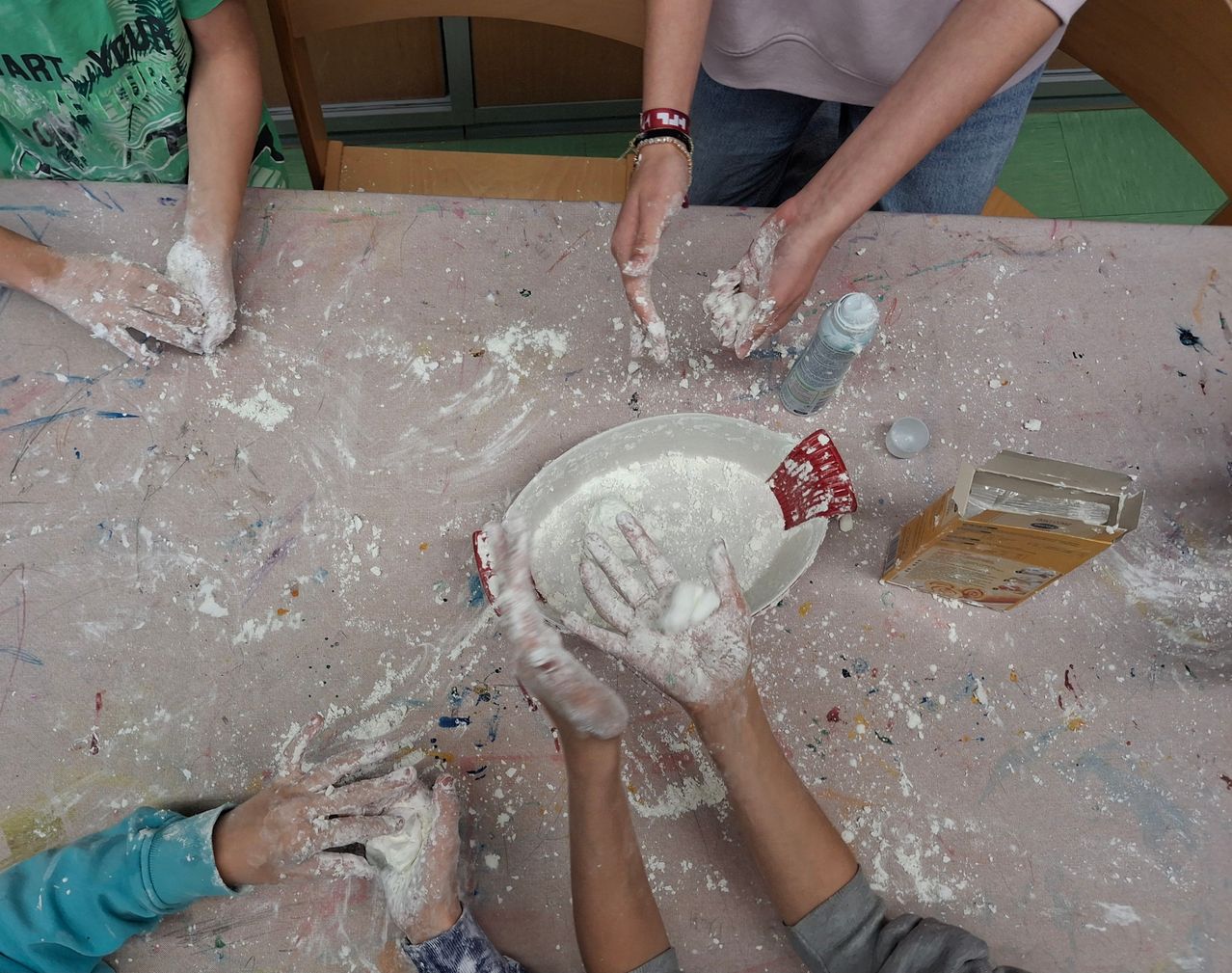 Eine Gruppe von Kindern sitzt um einen mit weißem Pulver bedeckten Tisch. Sie haben ihre Hände mit weißem Pulver bedeckt, während sie sich auf das Malen vorbereiten. Auf dem Tisch befinden sich eine weiße Schüssel mit weißem Pulver, eine Schachtel und eine Sprühflasche.