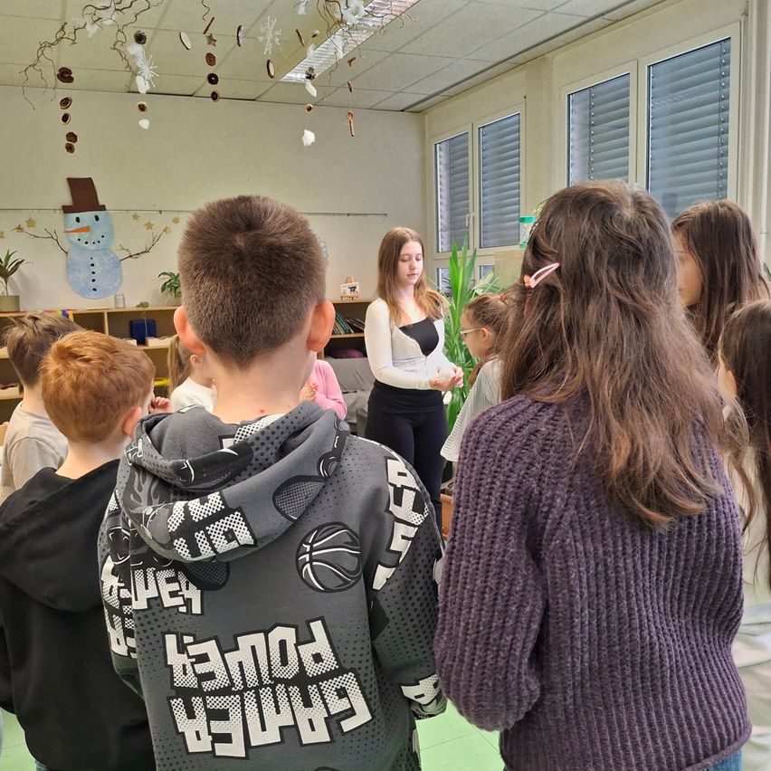 Ein Klassenzimmer mit einer Lehrerin, die den Schülern etwas erklärt. Einige Schüler tragen Kapuzenpullover, andere haben lange Haare. Das Klassenzimmer hat Bücherregale, Pflanzen und Dekorationen.