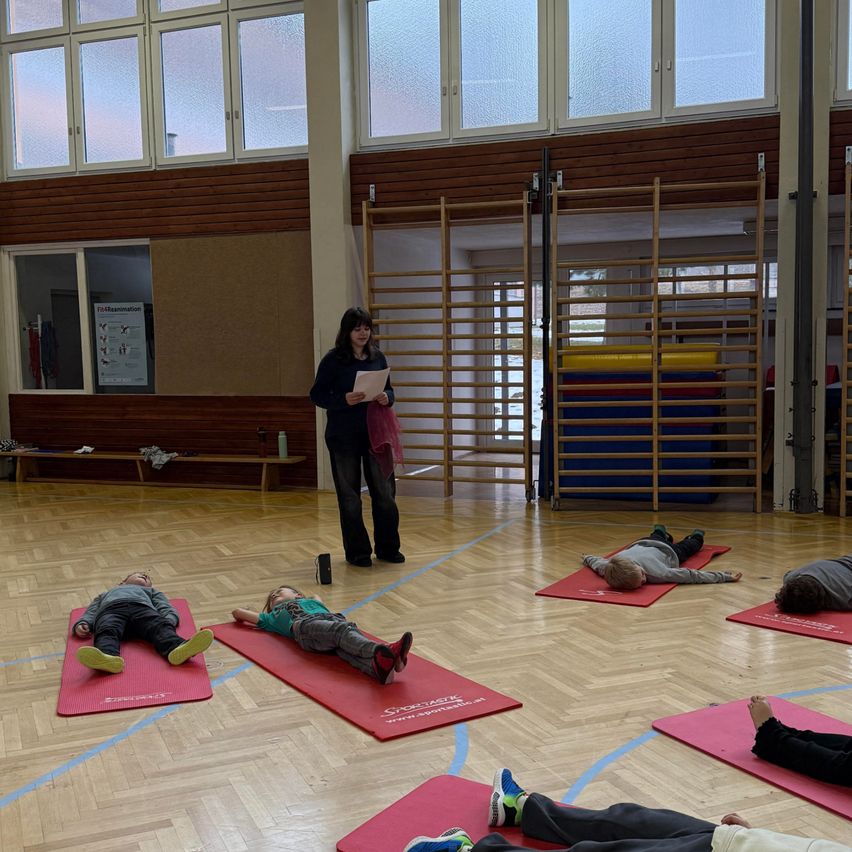In einer Turnhalle liest eine Frau Anweisungen von einem Papier, während mehrere Kinder auf roten Matten liegen, möglicherweise Yoga oder Entspannungsübungen machen. In der Nähe befinden sich Bänke und eine gelbe Wand.
